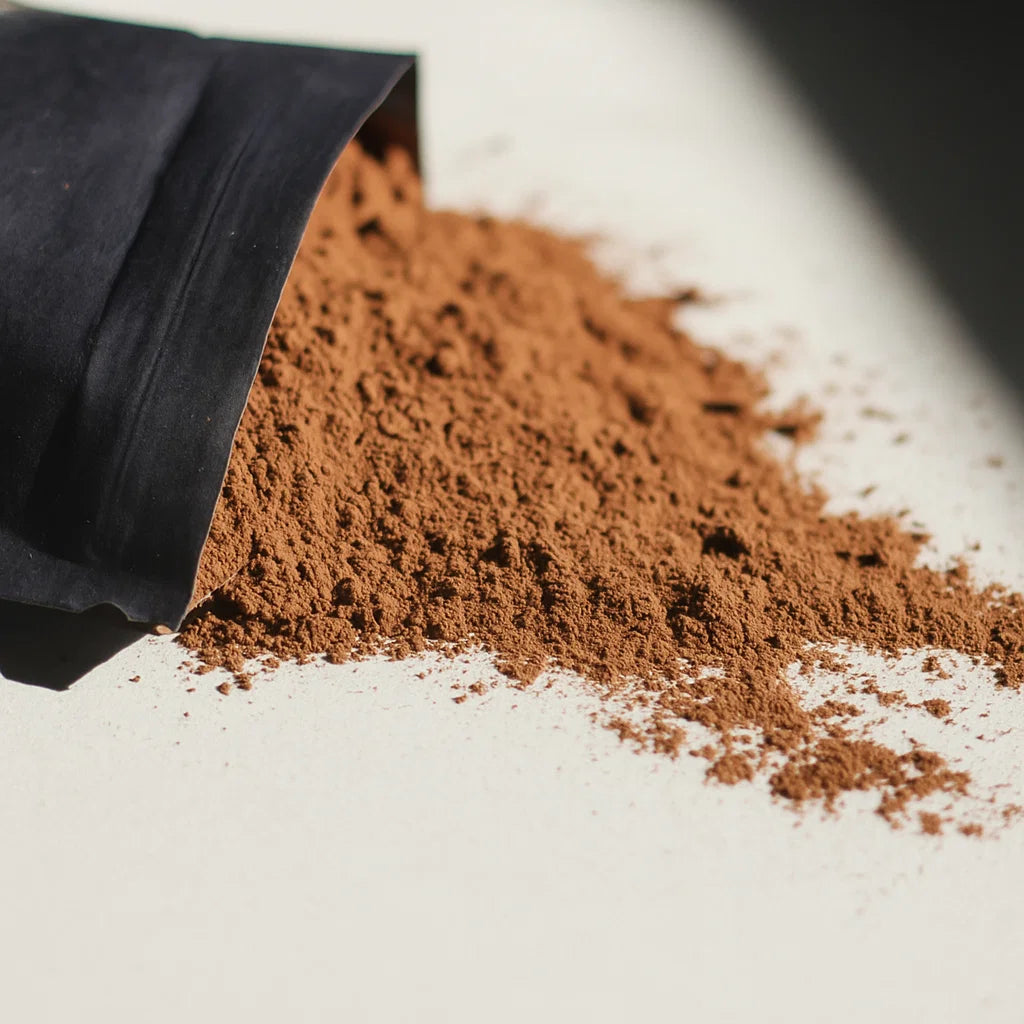 A black Shroomate Mushroom Hot Chocolate packet spills a mound of brown adaptogenic cacao onto a white surface, with scattered powder extending from the pile and illuminated by natural light.